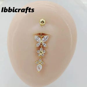 Puede incluir: Anillo para el ombligo dorado con diseño de mariposa y flores. La joya presenta piedras preciosas claras y brillantes y un dije colgante con forma de flor y lágrima. La bola superior y la mariposa son doradas.