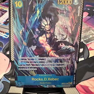 May include: A One Piece trading card featuring the character Rocks.D.Xebec with a cost of 10 and 12000 power. The card displays a dynamic illustration of the character in a battle stance with blue energy effects and includes detailed game text.