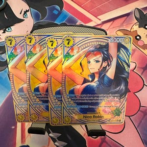 May include: Four trading cards featuring Nico Robin from the Straw Hat Crew. Each card has a gold background with a portrait of the character, the number 7, and game text. The cards are on a colourful playmat.