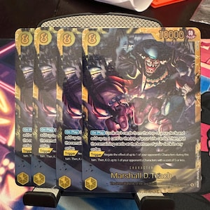 May include: Four identical Marshall D. Teach trading cards featuring a dark, illustrated character with a wide grin. The cards display a power level of 10000, yellow icons, and detailed game text instructions for On Play and Trigger abilities in English.