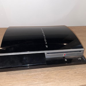 May include: A black Sony PlayStation 3 game console with a glossy finish. The top surface features the text PLAYSTATION 3 in silver lettering. The front panel includes a silver disc slot, a small PlayStation logo, and power and eject icons.