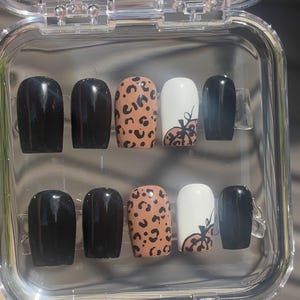 May include: A set of press-on nails in a clear plastic case. The nails feature a variety of designs, including solid black, leopard print, and white with a pumpkin design. The nails are oval-shaped.