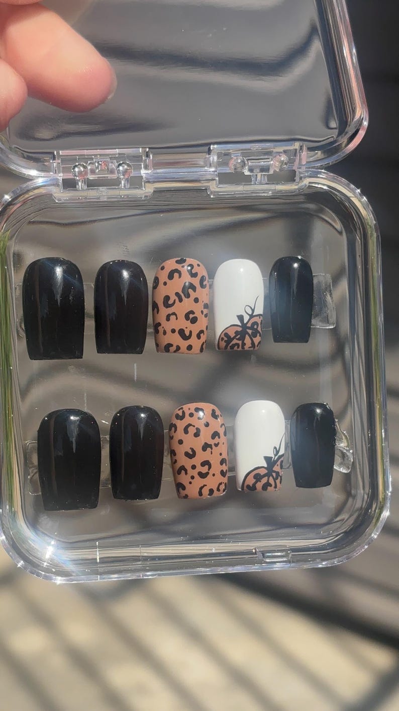 May include: A set of press-on nails in a clear plastic case. The nails feature a variety of designs, including solid black, leopard print, and white with a pumpkin design. The nails are oval-shaped.