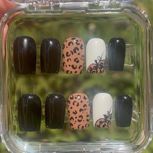 May include: A set of ten artificial nails in a clear plastic case. The nails feature a mix of solid black, leopard print, and white with a pumpkin design. The nails are oval-shaped and appear to be ready for application.