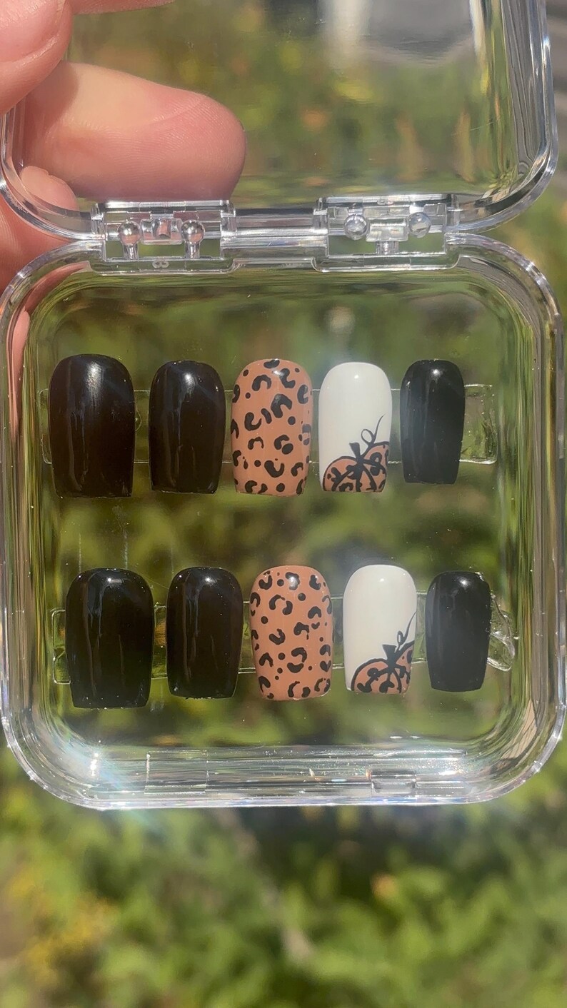 May include: A set of ten artificial nails in a clear plastic case. The nails feature a mix of solid black, leopard print, and white with a pumpkin design. The nails are oval-shaped and appear to be ready for application.