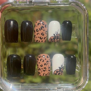 May include: A set of press-on nails in a clear plastic case. The nails feature a variety of designs, including solid black, leopard print, and white with a pumpkin design. The nails are a medium length and have a rounded shape.