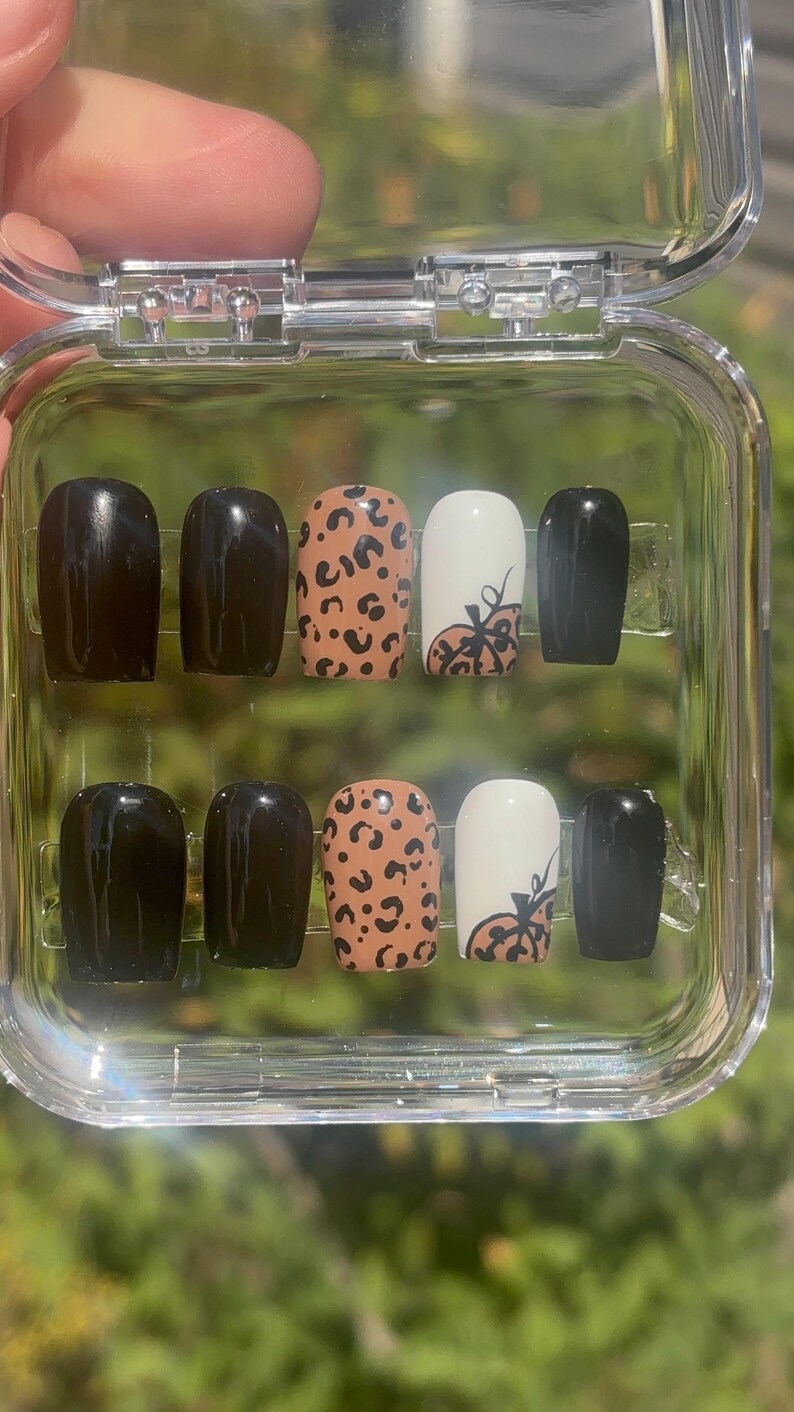 May include: A set of press-on nails in a clear plastic case. The nails feature a variety of designs, including solid black, leopard print, and white with a pumpkin design. The nails are a medium length and have a rounded shape.