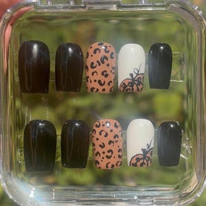 May include: A set of ten artificial nails in a clear plastic case. The nails feature a variety of designs, including solid black, solid brown, leopard print, and white with a pumpkin design. The nails are oval-shaped.