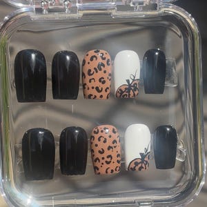 May include: A set of press-on nails in a clear plastic case. The nails feature a variety of designs, including solid black, leopard print, and white with a pumpkin design. The nails are oval-shaped.