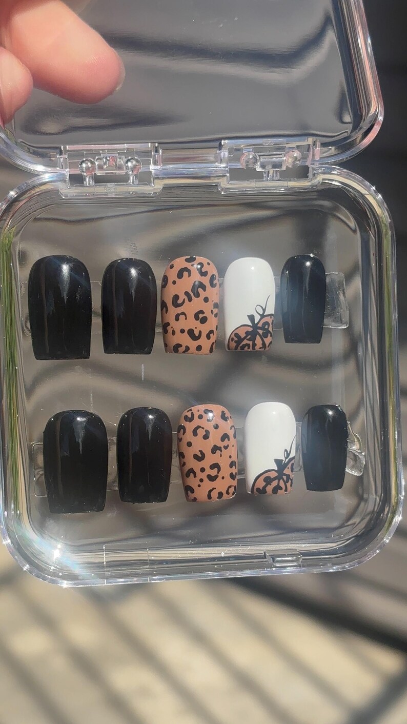May include: A set of press-on nails in a clear plastic case. The nails feature a variety of designs, including solid black, leopard print, and white with a pumpkin design. The nails are oval-shaped.