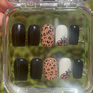 May include: A set of press-on nails in a clear plastic case. The nails feature a variety of designs, including solid black, leopard print, and white with a pumpkin design. The nails are a medium length and have a rounded shape.