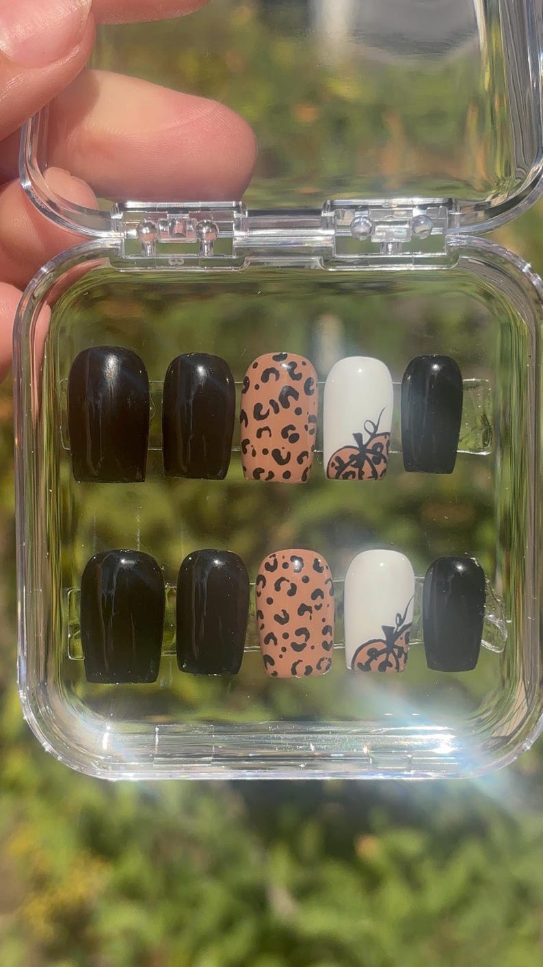 May include: A set of press-on nails in a clear plastic case. The nails feature a variety of designs, including solid black, leopard print, and white with a pumpkin design. The nails are a medium length and have a rounded shape.