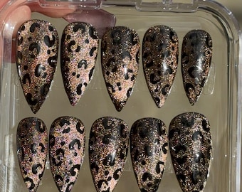 May include: A set of ten almond-shaped false nails with a leopard print design. The nails have a black leopard print pattern over a shimmering gold and pink base, presented in a clear plastic case.