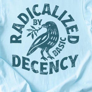 Puede incluir: Camiseta azul claro con mangas enrolladas. La camiseta presenta un gráfico verde oscuro de un pájaro con una ramita, rodeado por las palabras "RADICALIZED BY BASIC DECENCY". La camiseta está hecha de material suave.