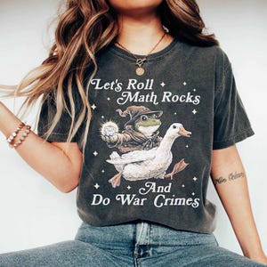 Puede incluir: Camiseta gris oscuro con el texto "Let's Roll Math Rocks And Do War Crimes". El diseño presenta una rana con sombrero de mago montada en un ganso blanco, sosteniendo un dado. La camiseta tiene un aspecto vintage.