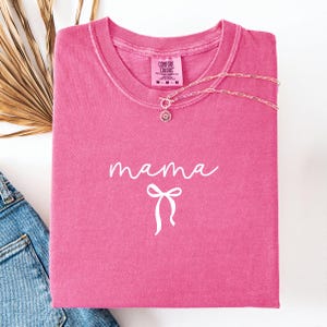 Peut inclure: Un t-shirt rose vif avec le mot "mama" en écriture blanche et un motif de nœud blanc. Un collier en argent avec un pendentif en forme de soleil est drapé sur le t-shirt. Le t-shirt est plié sur une surface blanche.