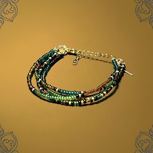 May include: A multi-strand beaded bracelet featuring green, brown, and black beads. The bracelet has a gold-colored clasp and chain for adjustable sizing. The beads are small and evenly spaced, creating a delicate and intricate design.