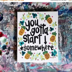 May include: A white canvas with a hand-painted pineapple design and the text "you gotta start somewhere". The background is decorated with pink, blue, and green flowers.