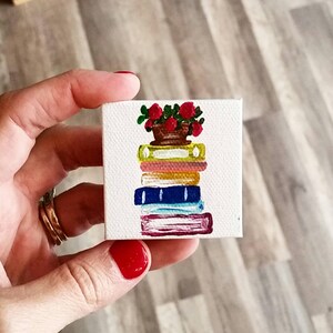 Op de afbeelding: Een miniatuurschilderij op canvas toont een stapel kleurrijke boeken, bekroond met een pot met rode rozen. De boeken zijn geschilderd in tinten geel, oranje, blauw en bordeaux. Het canvas wordt in een hand gehouden.