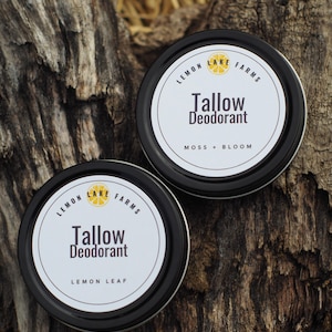 May include: Two round black metal tins of tallow deodorant with white labels. The labels feature the text Lemon Lake Farms, Tallow Deodorant, and the scents Moss + Bloom or Lemon Leaf, along with a small yellow lemon slice graphic icon.