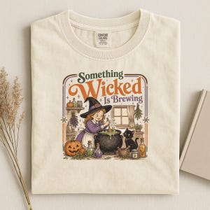 May include: A cream-colored t-shirt featuring a graphic of a young witch in a purple dress and black hat stirring a cauldron. The text reads "Something Wicked Is Brewing" above the illustration, which includes a black cat, pumpkin, and bottles.