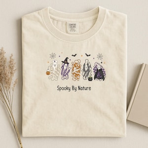 May include: A cream-colored t-shirt featuring a graphic of five ghosts in various costumes, including a nurse, a witch, a mummy, and a vampire. The design includes spiderwebs, bats, stars, and the text Spooky By Nature printed in black ink.