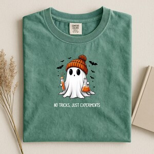 May include: A sage green crewneck t-shirt featuring a graphic of a white ghost wearing an orange knit beanie. The ghost holds a test tube and a flask filled with orange liquid, surrounded by small black bats. The text reads: NO TRICKS, JUST EXPERIMENTS.