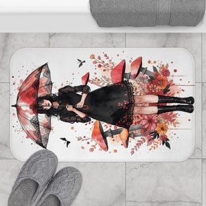 Könnte beinhalten: Rechteckige Badematte mit einer Aquarellillustration einer Person in einem schwarzen Kleid, die einen roten Regenschirm hält. Das Design umfasst florale Elemente und rote Schuhe. Graue Hausschuhe sind im Vordergrund.
