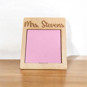 Puede incluir: Un portamemos de madera con el nombre "Mrs. Stevens" grabado en la parte superior. El soporte tiene una abertura rectangular con un bloc de notas rosa en el interior. La madera tiene un acabado claro y natural.