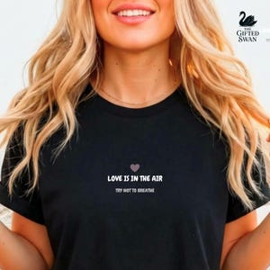 Op de afbeelding: Zwart T-shirt met de tekst "LOVE IS IN THE AIR" boven "TRY NOT TO BREATHE" in het wit. Een klein paars hartje staat boven de tekst. Het T-shirt wordt gedragen door een persoon met lang blond haar.