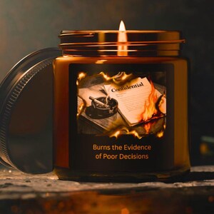 May include: A lit candle in a brown glass jar with a metal lid. The label features a graphic of a burning document labeled Confidential and an ashtray with a cigarette, with the text Burns the Evidence of Poor Decisions printed below.
