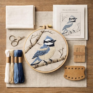 Op de afbeelding: Een borduurpakket met een blauwe gaai-ontwerp. De set bevat een borduurring, stof, garen in verschillende tinten blauw en crème, een schaar, naalden en een naaldhouder van kunstleer. De voltooide borduursel toont een blauwe gaai op een tak.