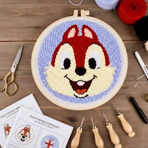 May include: A punch needle embroidery kit with a cartoon character's face in red, cream, and black. The design is set in a wooden hoop with a light blue background. The kit includes needles, scissors, and instructions.