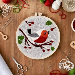 May include: An embroidered hoop art piece depicting a red, white, and grey bird on a brown branch with green leaves and red berries. The bird is set against a grey background. Craft supplies, including scissors and needles, surround the hoop.