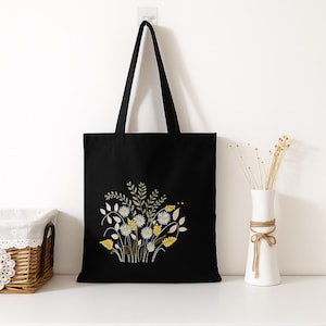 Peut inclure: Sac fourre-tout noir avec un motif floral brodé. Le motif comprend des fleurs blanches et jaunes, des feuilles vertes et des tiges marron. Le sac a des anses noires et est fabriqué dans un tissu résistant. Idéal pour les courses.