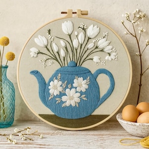 Könnte beinhalten: Gestickte Wandkunst mit einer blauen Teekanne mit weißen Blumenakzenten und einem Strauß weißer Blumen. Das Kunstwerk ist in einem hölzernen Stickrahmen gefasst und schafft eine charmante, handgefertigte Ästhetik. Der Hintergrund ist ein neutrales Beige.