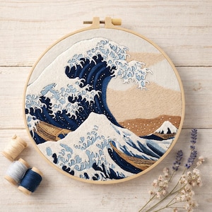 May include: Embroidered artwork of a large ocean wave in shades of blue, white, and tan, set in a wooden hoop. The design includes a mountain and boats. Spools of thread and flowers are arranged around the hoop.