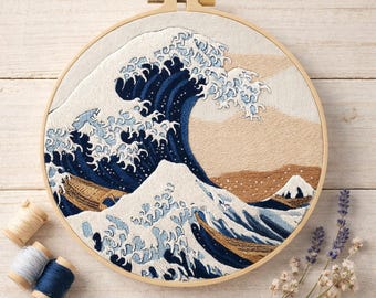May include: Embroidered artwork of a large ocean wave in shades of blue, white, and tan, set in a wooden hoop. The design includes a mountain and boats. Spools of thread and flowers are arranged around the hoop.