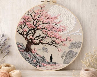 May include: Embroidered artwork in a wooden hoop. The design features a cherry blossom tree with pink flowers, a full moon, and a figure standing near a rocky landscape. The color palette includes pink, brown, gray, and cream.