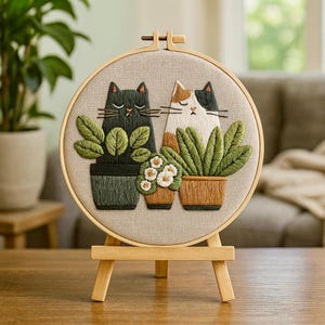 May include: Embroidered artwork showing two cats with closed eyes, each next to a potted plant. The cats are black and calico, with green and brown plants. The artwork is displayed on a wooden easel.