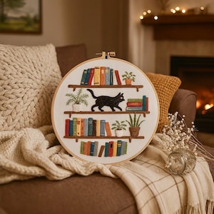 Könnte beinhalten: Gestickter Stickrahmen mit einer schwarzen Katze auf einem Bücherregal mit bunten Büchern und Pflanzen. Der weiße Rahmen hat einen Holzrahmen, ideal für die Heimdekoration. Ein charmantes Textilkunstwerk.