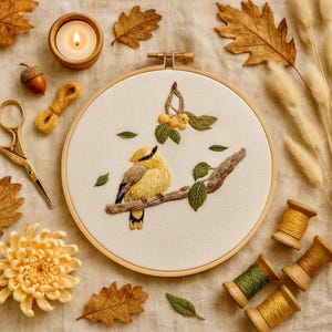 Puede incluir: Una detallada pieza de arte de bordado en bastidor que representa un pájaro amarillo posado en una rama con hojas verdes. La obra de arte está rodeada de elementos temáticos de otoño como hojas, una vela y carretes de hilo. El pájaro es el punto focal.