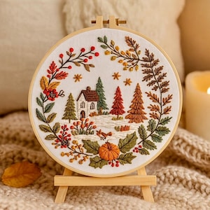 May include: An embroidered hoop art piece depicting an autumnal scene. The design shows a house, trees, a pumpkin, and a fox, framed by fall leaves in red, orange, and green hues. The hoop is displayed on a small wooden easel.