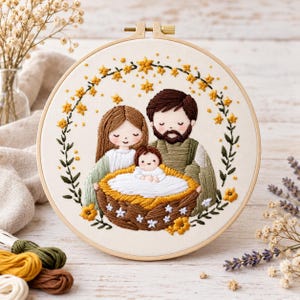 May include: A circular embroidery hoop featuring a hand-stitched scene of a family of three. A parent with long brown hair and a parent with a brown beard stand behind a baby in a woven basket, all surrounded by a wreath of yellow flowers.