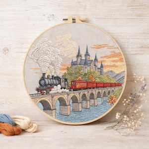May include: Embroidered train scene in a wooden hoop. The train is black and red, crossing a stone bridge over water. A castle is in the background, with a sunset sky. Embroidery threads in blue, cream, and brown are in the lower left.