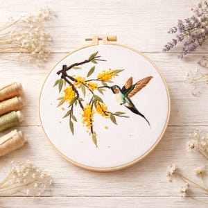 May include: A circular wooden embroidery hoop containing white fabric with a hand-stitched design of a hummingbird hovering near a branch with green leaves and small yellow flowers. The bird features brown, green, and white thread details.