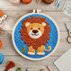 May include: A completed punch needle embroidery of a cartoon lion in a wooden hoop. The lion has a brown mane and tan face, set against a blue background. Craft supplies, including thread and scissors, are arranged around the hoop, ready for crafting.