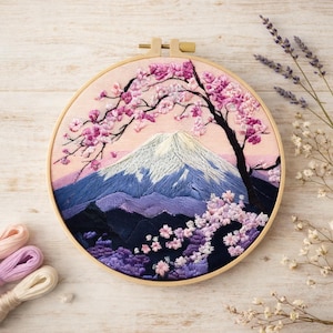 May include: A circular wooden embroidery hoop containing a fabric panel with a hand-stitched landscape. The design features a snow-capped mountain peak in shades of blue and white, framed by a dark tree with vibrant pink cherry blossom flowers.