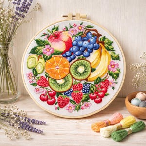 May include: Embroidered fruit design in a wooden hoop. The artwork features a vibrant arrangement of an apple, grapes, bananas, kiwi, orange slices, strawberries, and cherries. Pink flowers and green leaves surround the fruit. Embroidery floss and a small wooden bowl are also in the image.