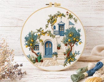 May include: An embroidered hoop art piece featuring a white house with blue doors and windows, surrounded by lemon trees. The artwork is set against a white wooden background, with embroidery floss in the foreground.